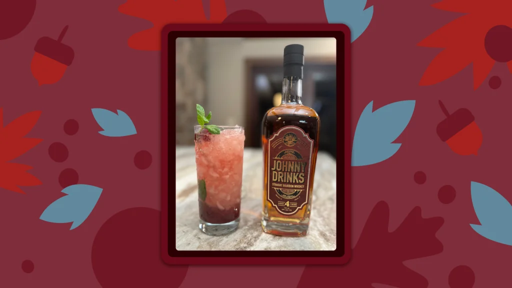 An icy cran-bourbon cocktail in a Tom Collins glass next to a Johnny Drinks bottle of bourbon.