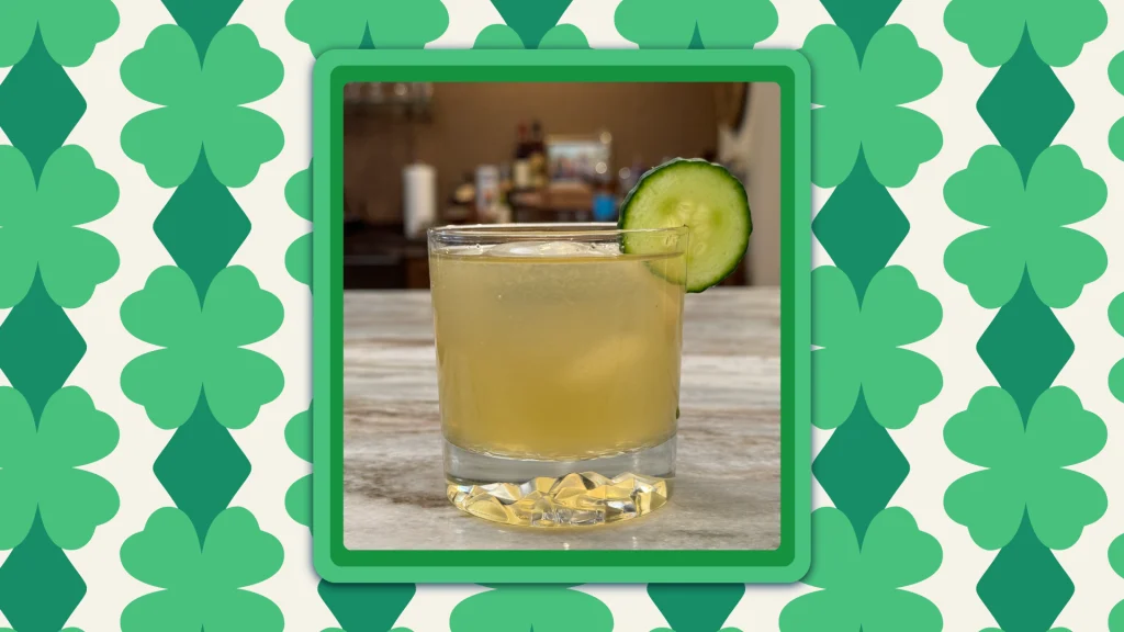 A glass featuring The Irish Maid cocktail, with a cucumber on the lip, on a marble bar top.